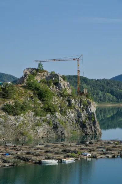 Vista matutina del Lago Zaovine con una grúa sobre una roca y piscifactorías flotantes en primer plano.