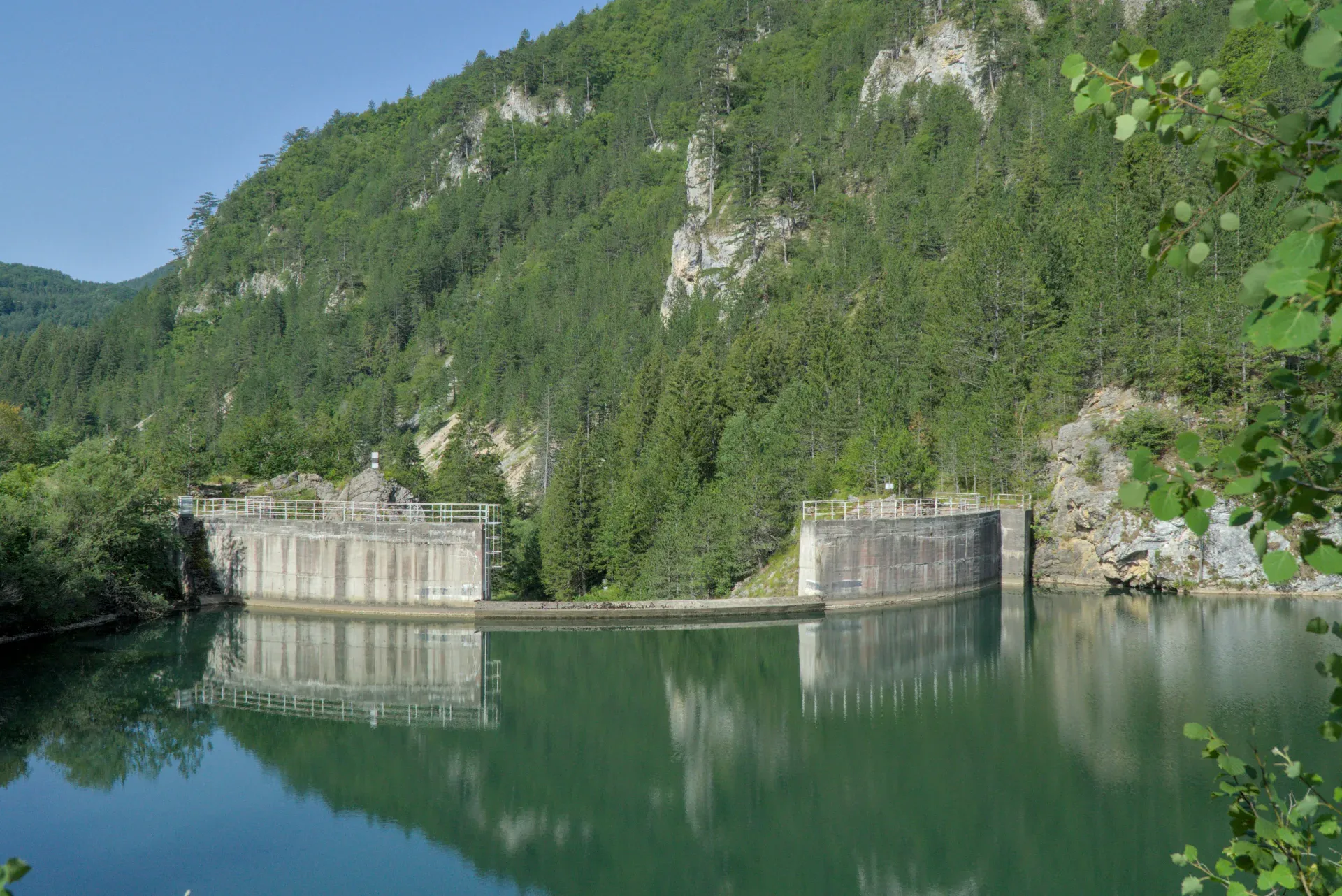 Spajići Lake featuring its concrete dam structure, reflected in green waters against forested hills.