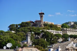 Towers and buildings on a green hillside in Genoa, Italy, under a clear blue sky.
