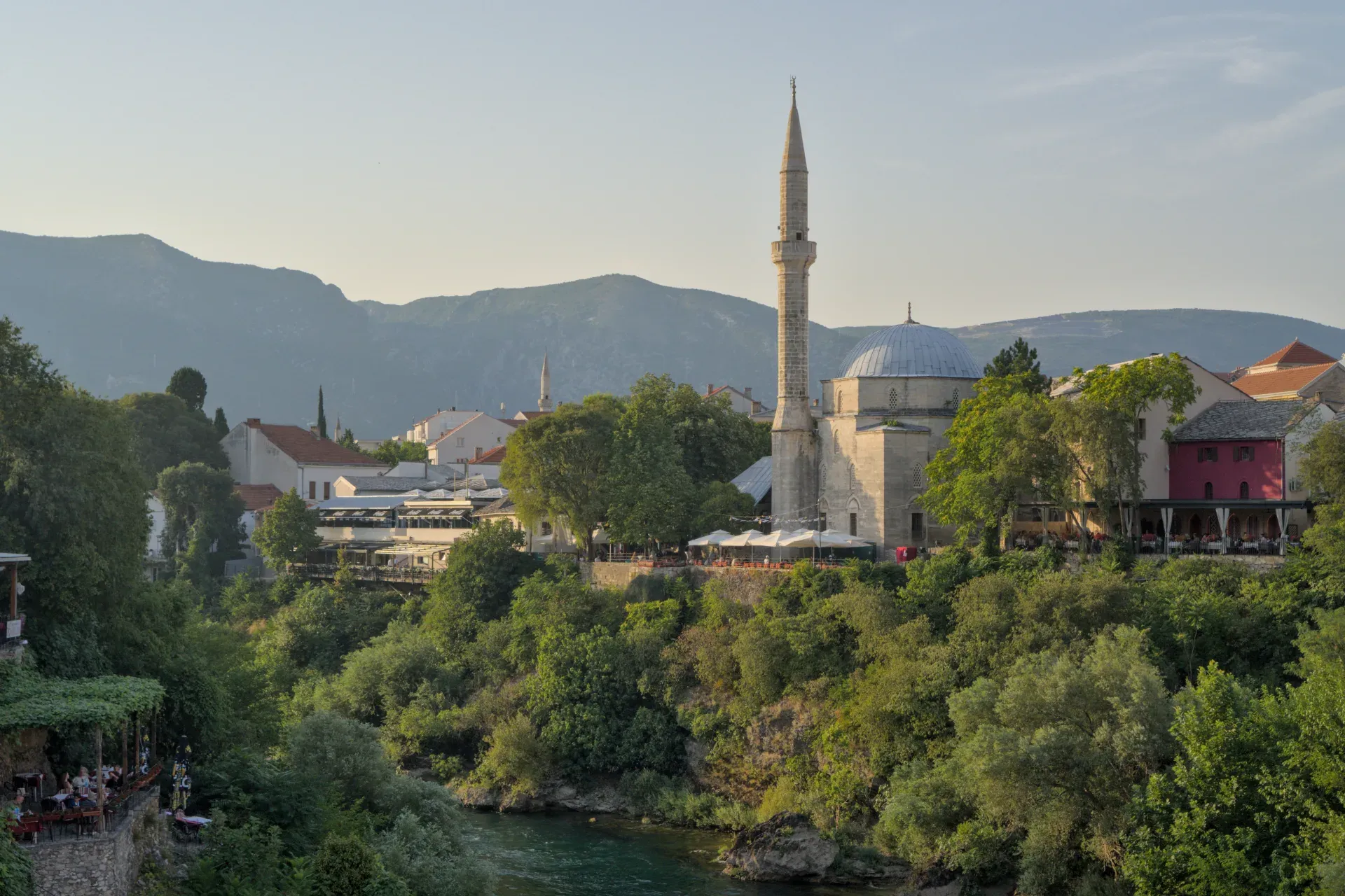 Golden hour landscape photograph of Mostar's old town featuring a mosque and lush greenery.