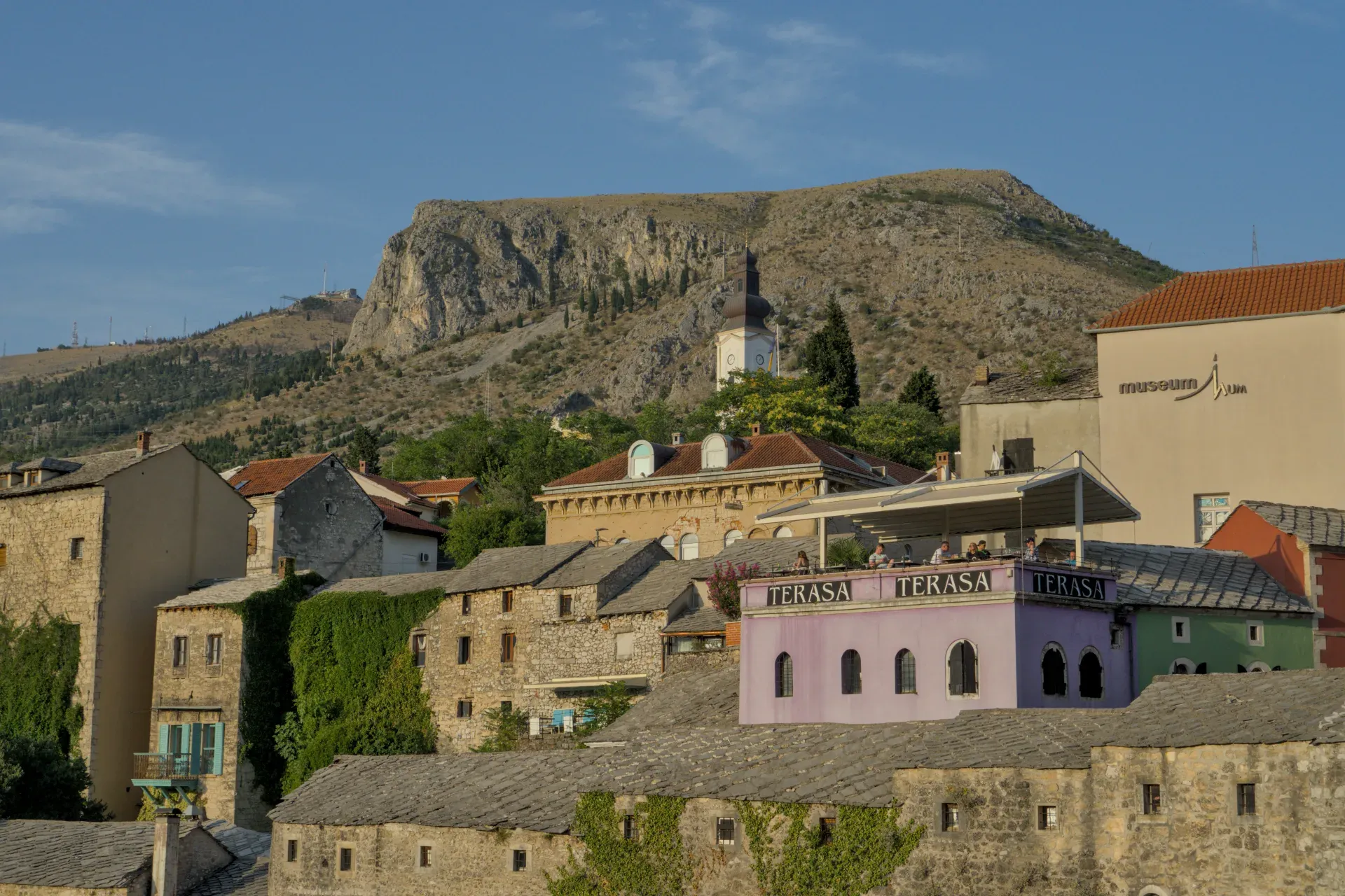 Panoramic landscape of Mostar's old town at dusk, featuring stone buildings, greenery, and a rocky mountain backdrop.