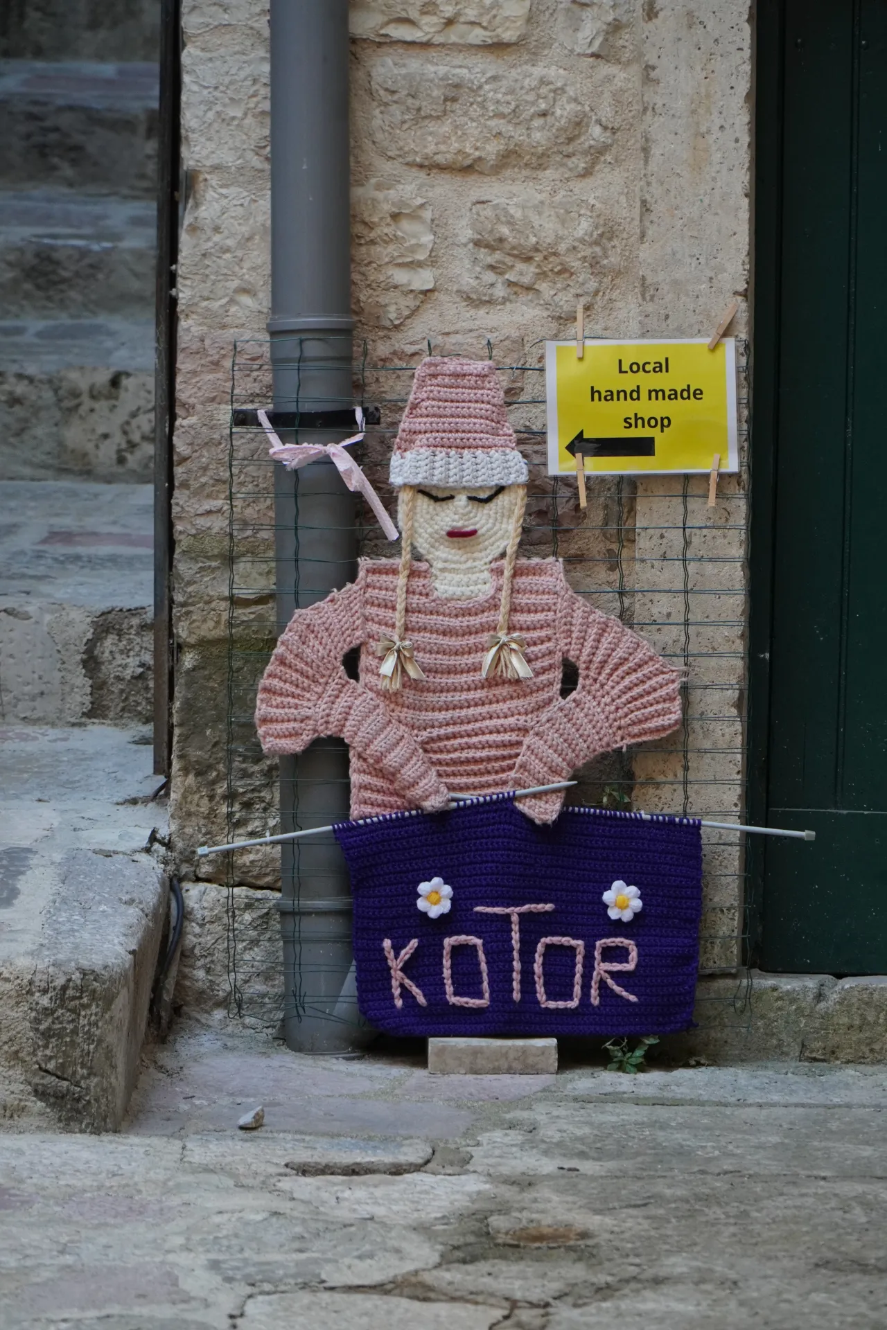 A knitted doll with a pink hat and sweater holding a purple sign that says 'Kotor' with small white flowers, in front of a stone wall in Kotor, Montenegro. A yellow sign pointing left advertises a 'Local hand made shop'.