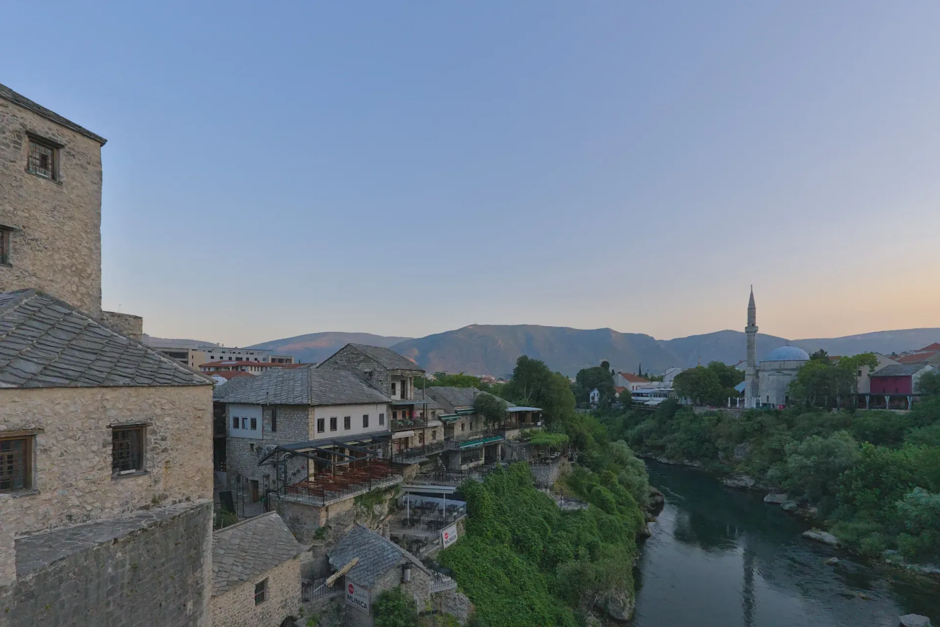 Panoramic view of Mostar's old town, featuring the Old Bridge, Neretva River, and a mosque in the distance.
