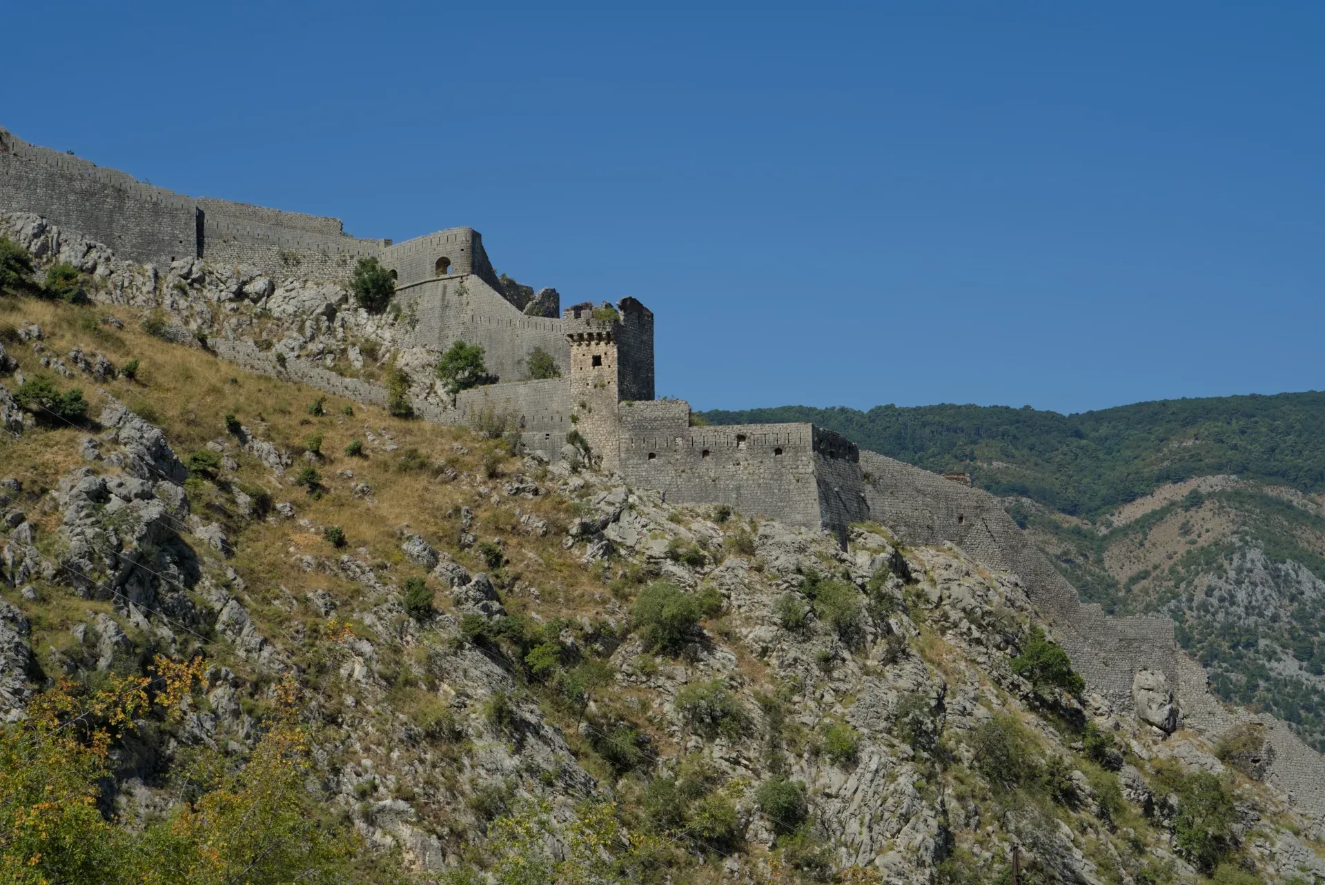 The старинные крепостные стены на скале of Kotor, Montenegro, stand majestically against a clear blue sky.