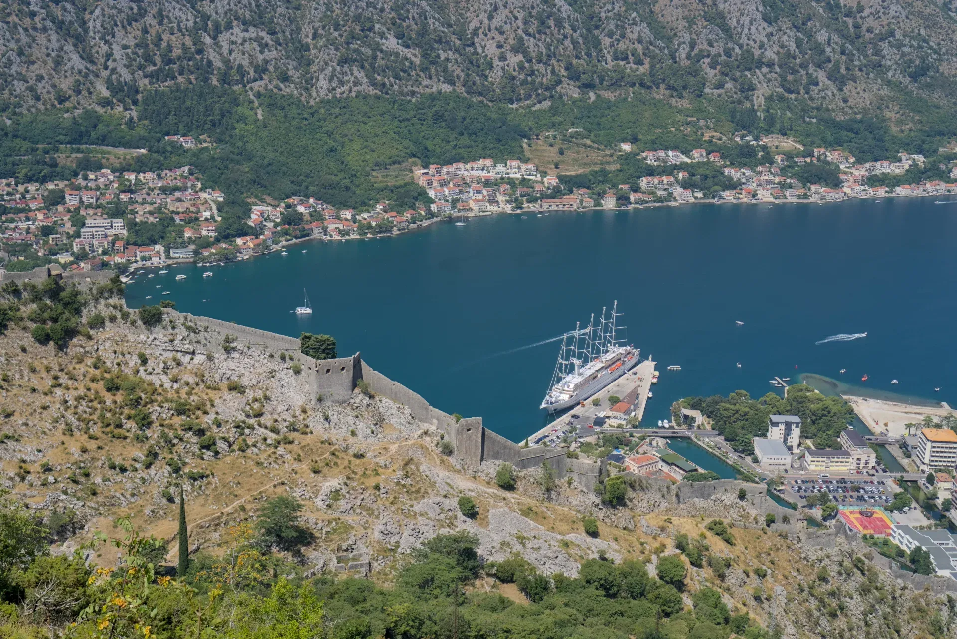 A massive sailing ship is docked in Kotor's harbor, Montenegro, with ancient fortress walls overlooking the scene.