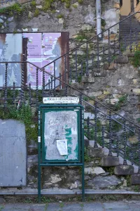 An old stone staircase with a decorative metal railing ascends alongside a stone wall. In the foreground, a green notice board displays weathered posters, and overgrown plants climb the wall and stairs.