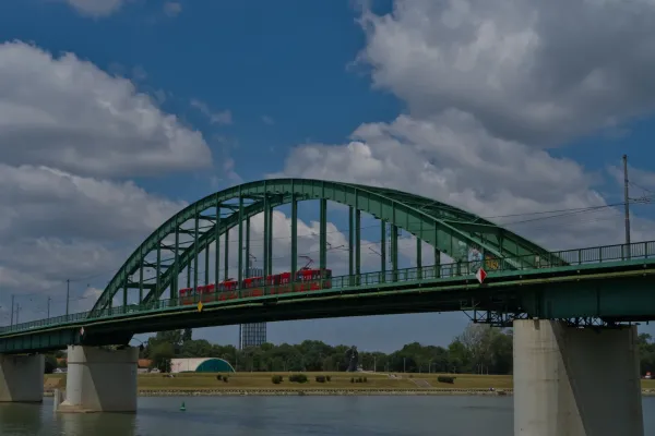 Stari Savski most u Beogradu sa crvenim tramvajem koji prolazi preko njega na pozadini plavog neba.