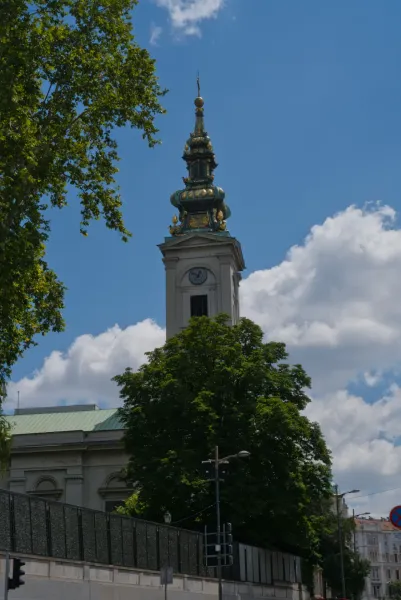 Eleganter Kirchturm in Belgrad vor blauem Himmel mit Wolken, umrahmt von Bäumen.
