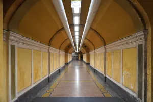 A long, symmetrical tunnel with yellow walls, white decorative trim, a patterned marble floor, and bright overhead fluorescent lighting.