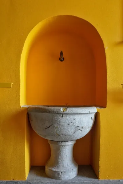 Un lavabo de piedra vintage se encuentra en un nicho arqueado de color amarillo brillante con un pequeño grifo de latón encima, contra una pared amarilla texturizada. El suelo está revestido de baldosas grises apagadas.