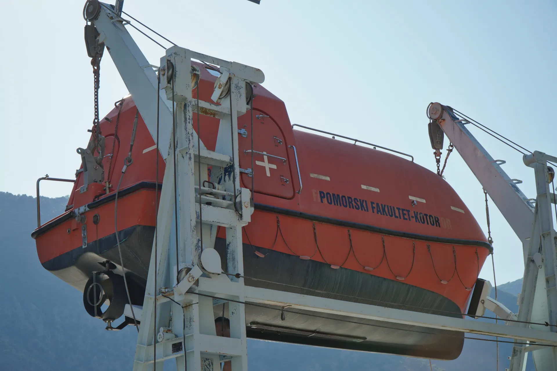 An orange lifeboat with 'POMORSKI FAKULTET-KOTOR' written on its side is hoisted by a crane, with a clear sky and distant mountains in the background.