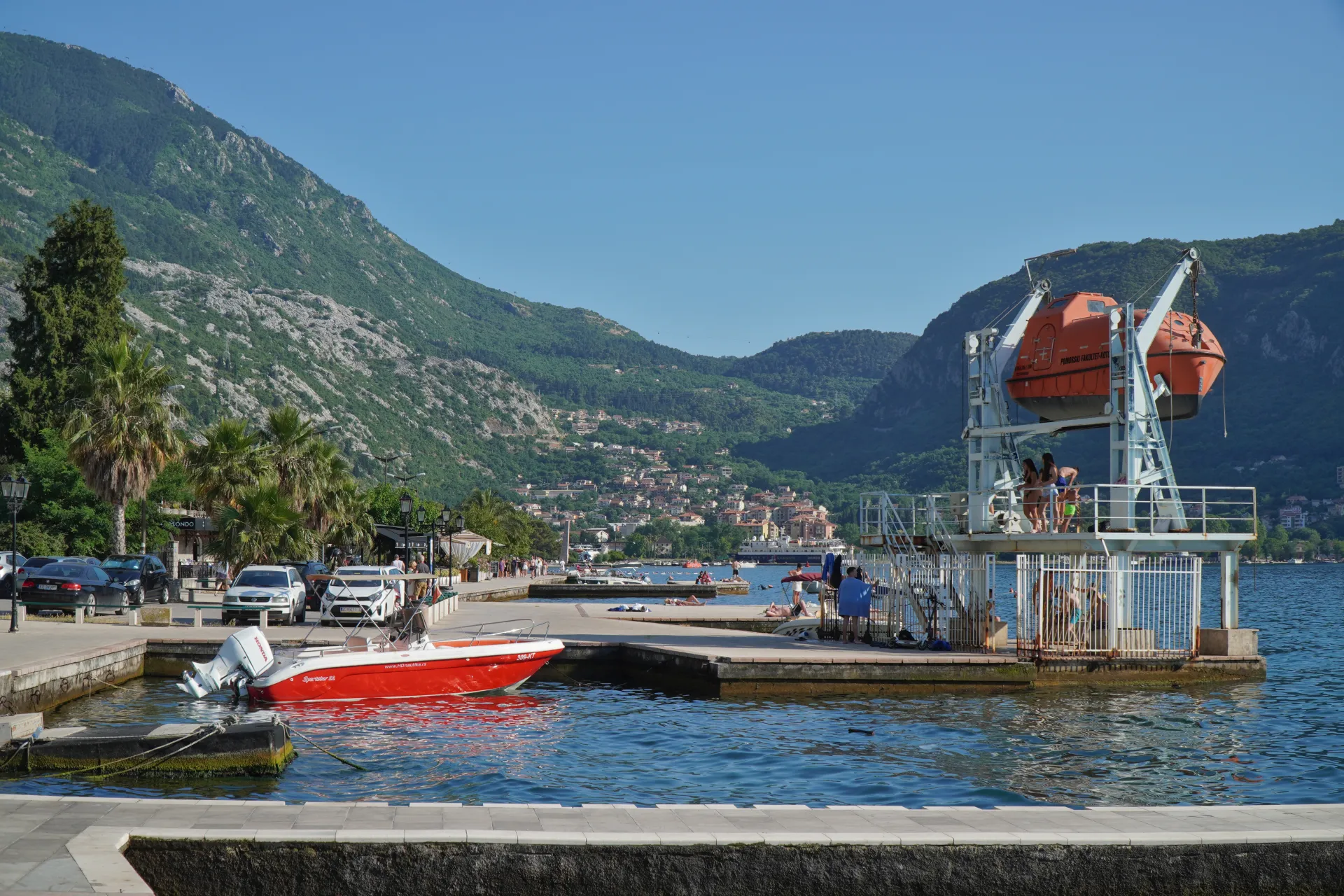 View of the Kotor Bay waterfront with a red speedboat in the marina, a diving platform with people, and lush green mountains dotted with houses in the background.