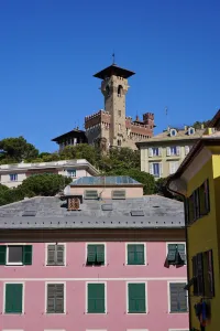 A historic castle with a prominent tower stands on a green hill, with various colorful residential buildings and rooftops in the foreground, under a clear blue sky in Genoa, Italy.