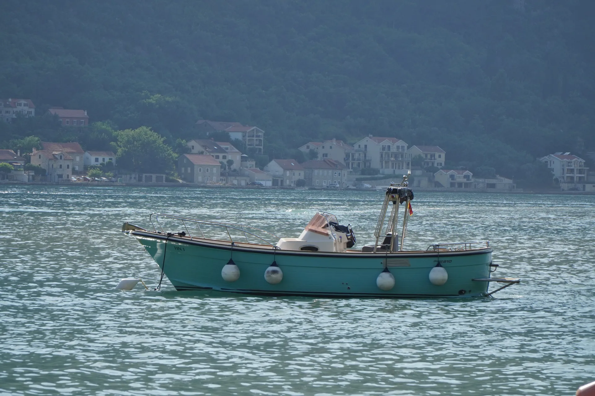 A teal-colored boat floats on the rippling turquoise waters of Kotor Bay, Montenegro. Buildings and lush green hills are visible on the shore in the distance.