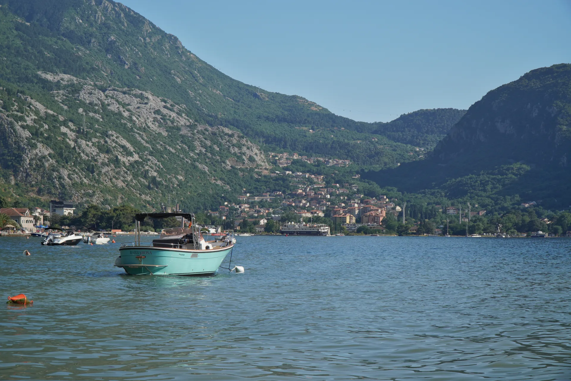 A turquoise boat named BARBAROSA floats on the calm, blue waters of Kotor Bay, Montenegro, with a charming town and green mountains rising behind it.
