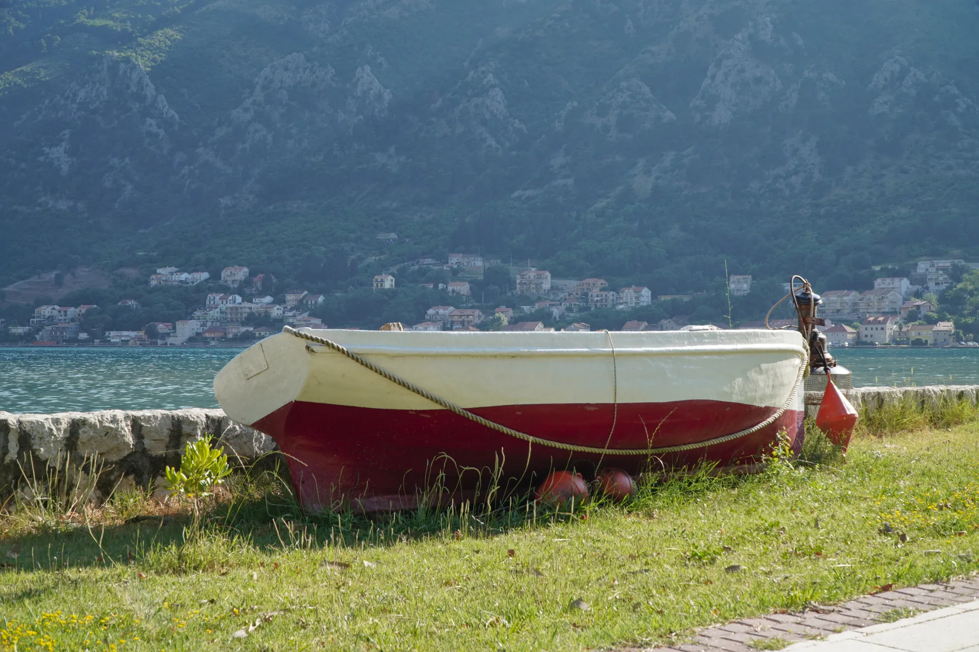 A red and white boat is pulled ashore on a grassy bank in Kotor, Montenegro, with the blue water of the Bay of Kotor and a hillside town in the background.