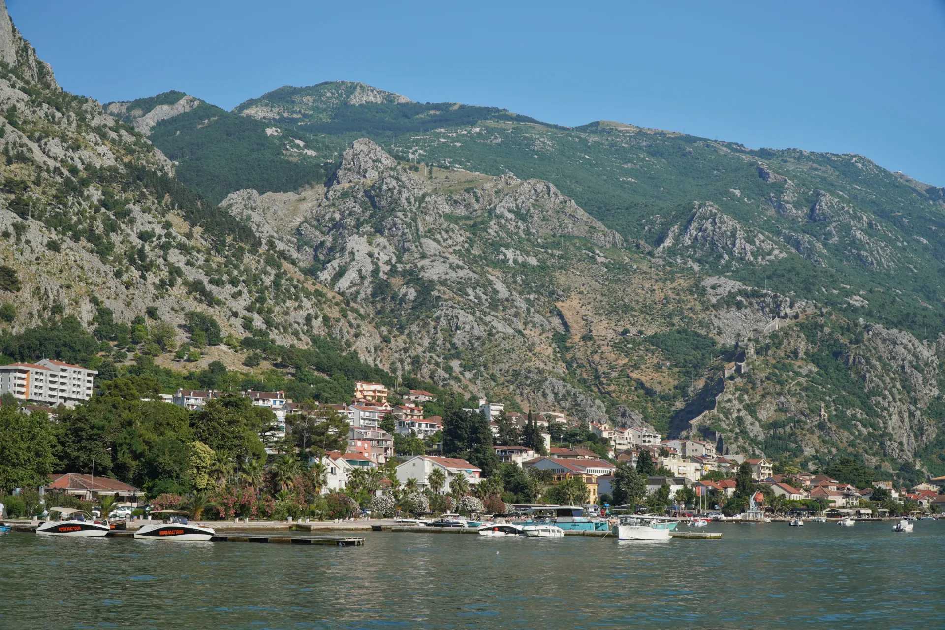 View of the Bay of Kotor with a town situated at the base of lush green mountains, boats are moored in the foreground on the water.