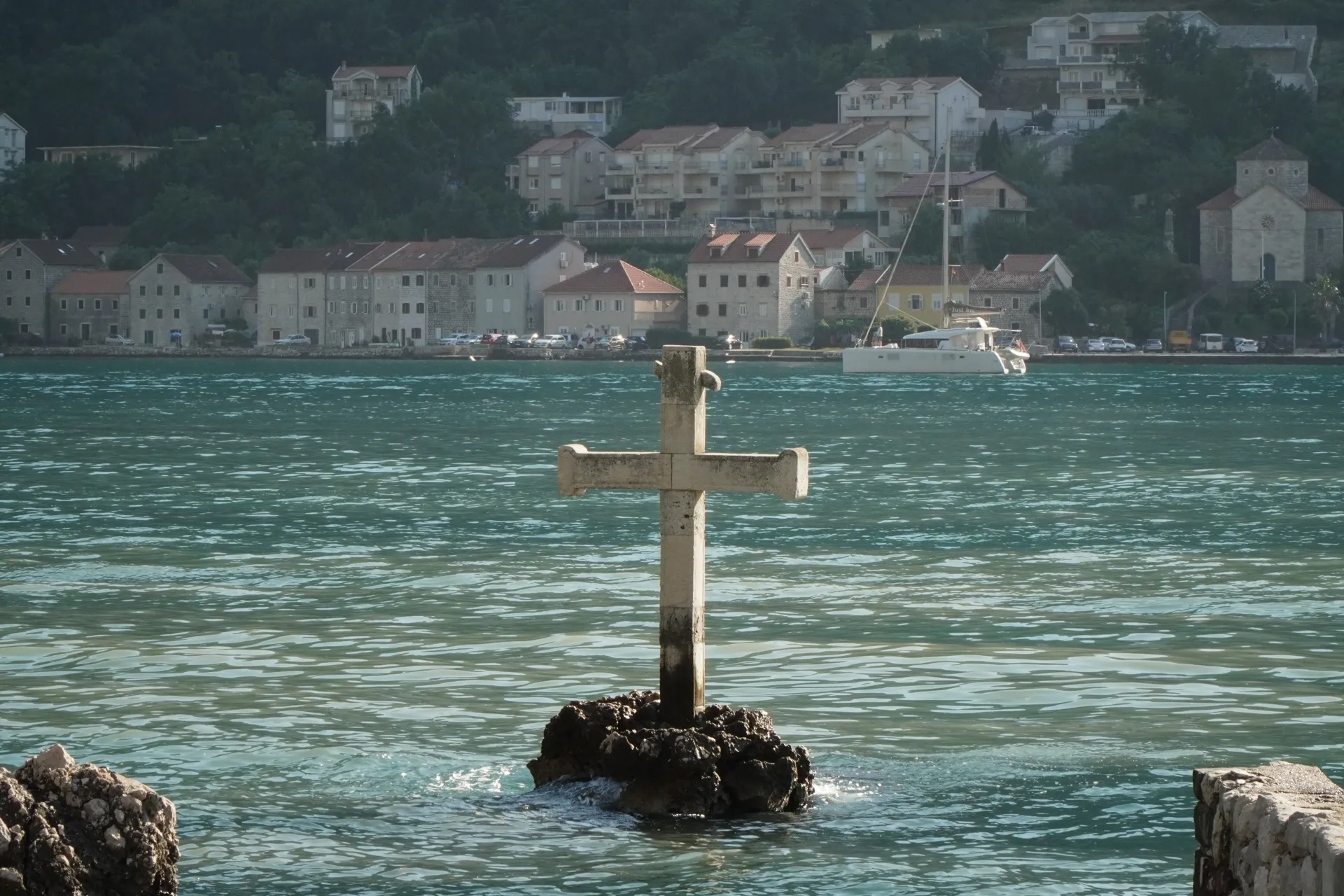 Kameni krst na steni u zalivu, sa zgradama i brdima u pozadini.