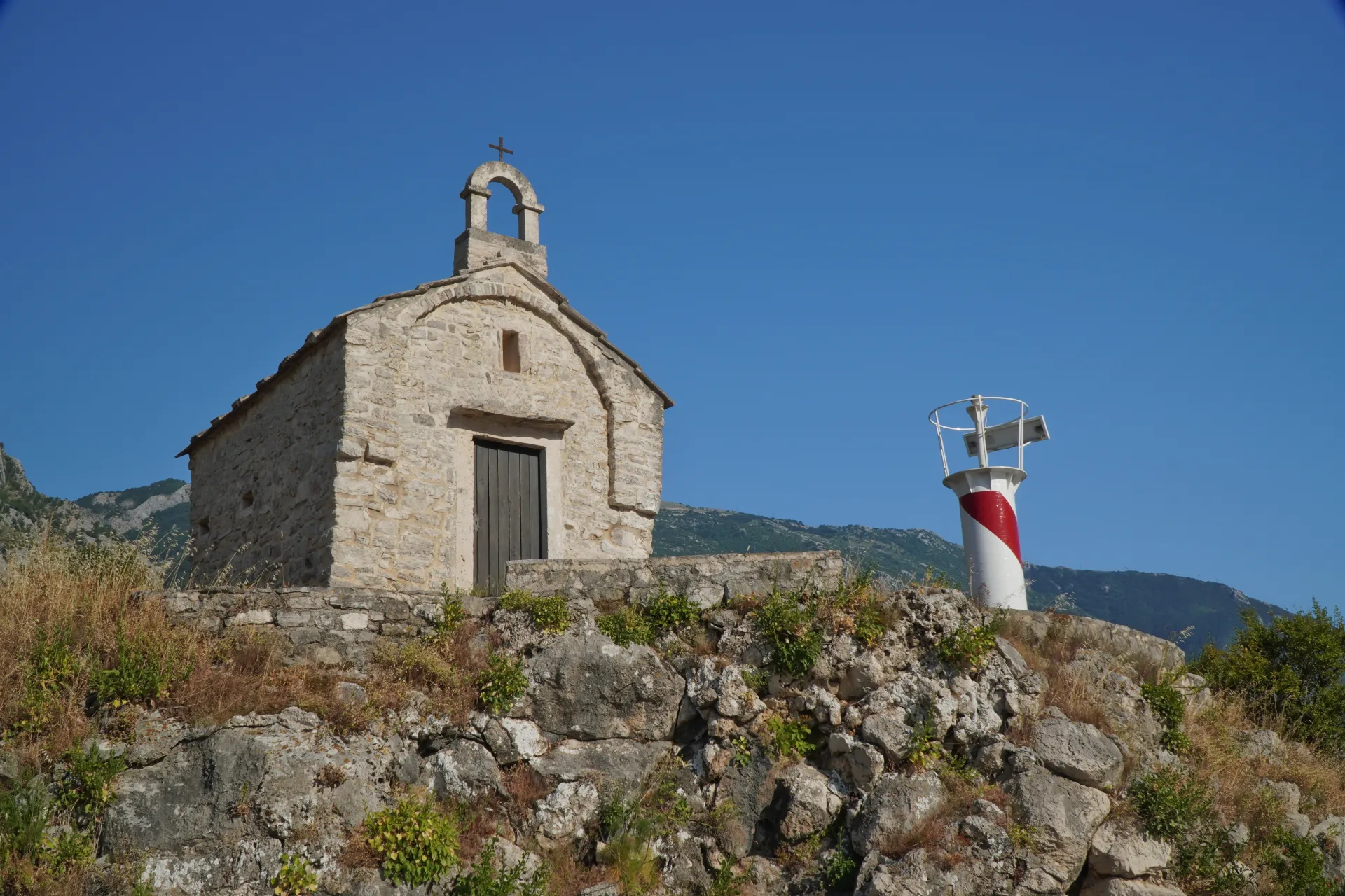 A small, ancient stone church with a bell tower and a cross, stands on a rocky hill next to a red and white striped lighthouse under a clear blue sky in Kotor, Montenegro.