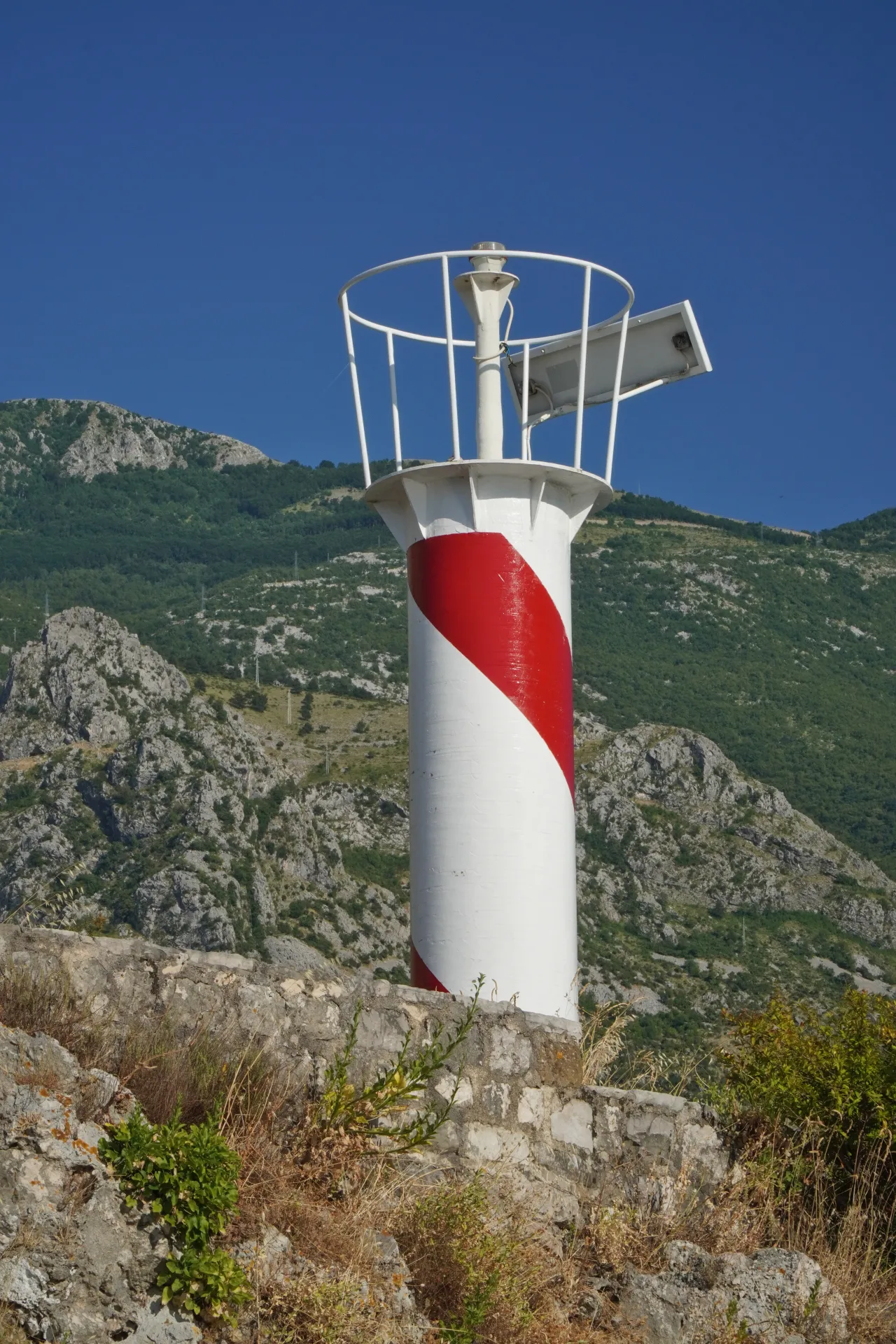 A red and white striped lighthouse in Kotor, Montenegro, with a clear blue sky and green mountains in the background.