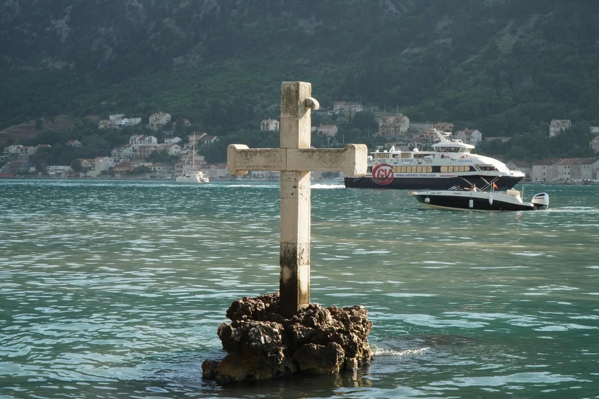 Kameni krst u vodi Kotorskog zaliva, Crna Gora, sa feribotom i gliserom u pozadini.