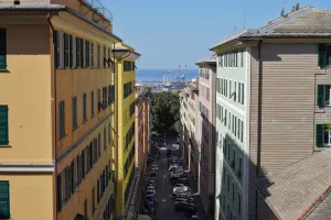 View down a narrow street in Genoa, Italy, flanked by colorful buildings with green shutters, leading to a distant view of the city's port with cranes and the blue sea.
