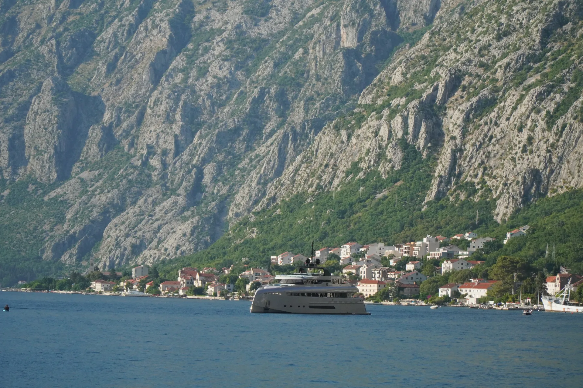 Elegancna, moderna superjahta klizi preko tamnoplave vode Kotorskog zaliva, sa šarmantnim gradom belih i crvenih krovova koji se prostire obalom, iza kojeg se nazire strma, kamena planina prekrivena zelenilom.