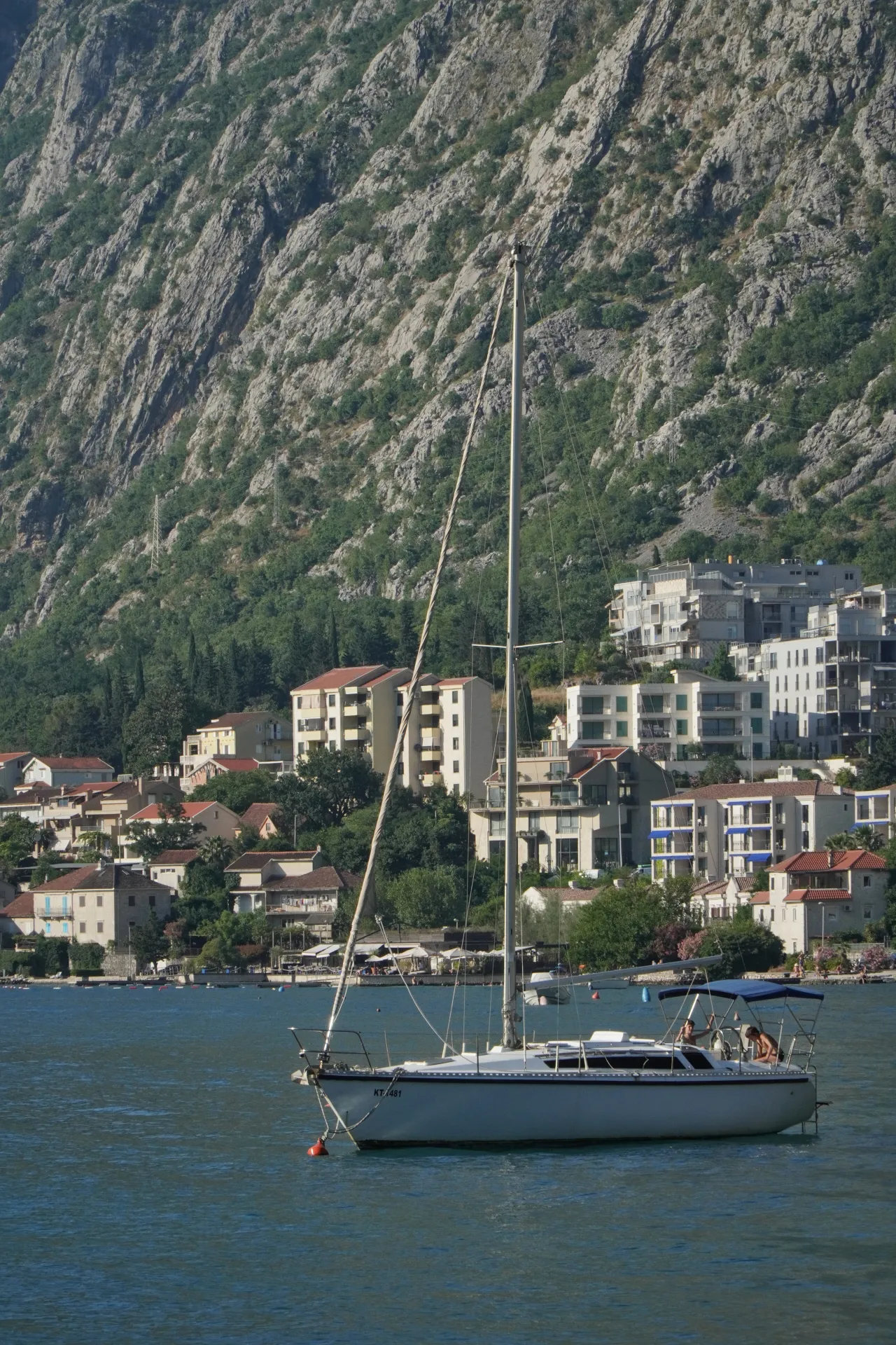 A white sailboat with a tall mast is anchored in the blue water of Kotor Bay, Montenegro. Behind the boat, a hillside town with red-tiled roofs and modern buildings slopes up to a rugged, green-covered mountain range.