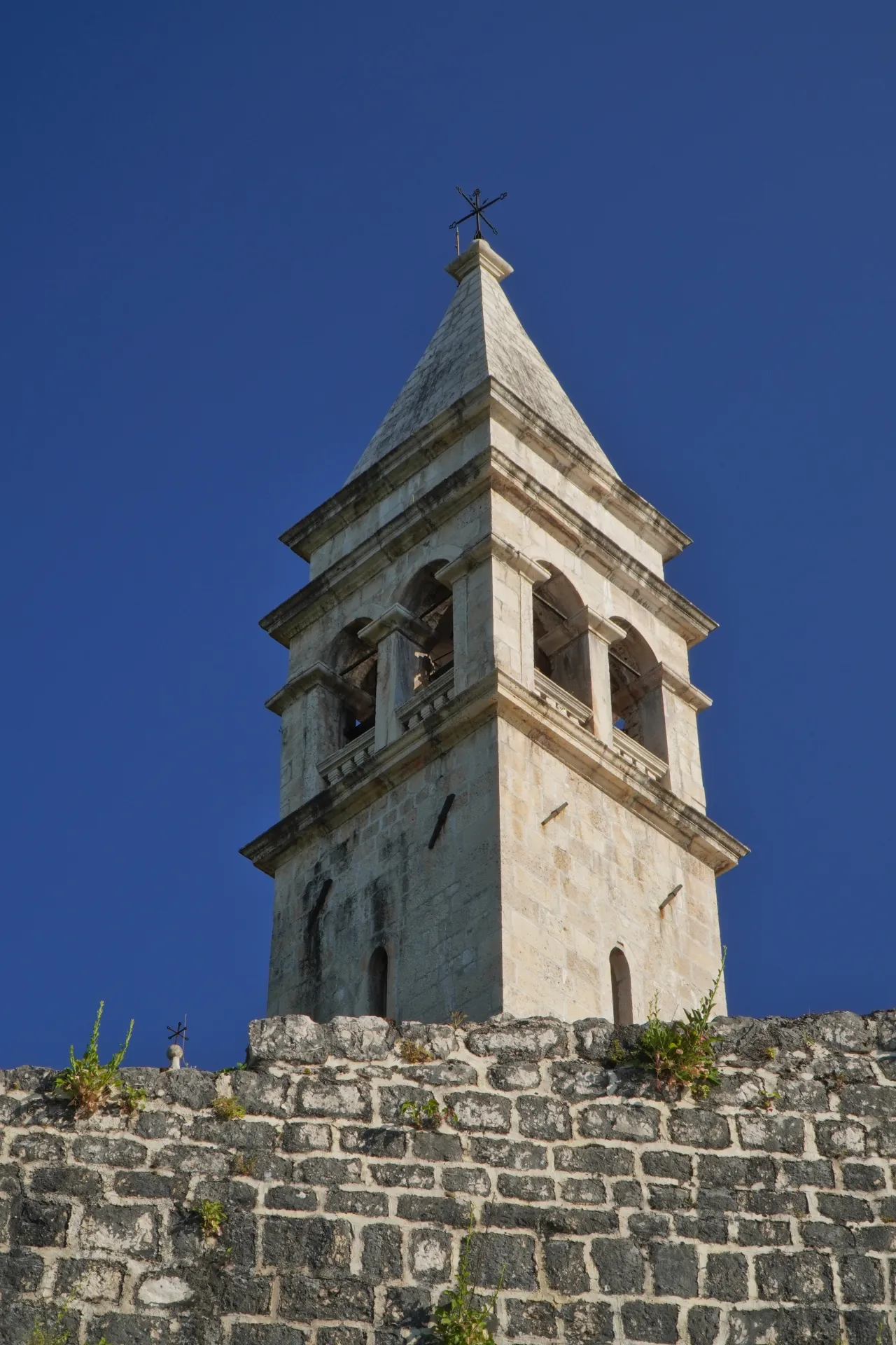 A close-up view of a weathered stone bell tower with arched openings, topped with a cross, rising against a deep blue sky. The tower is situated on top of an old, dark stone wall with some green plants growing on it.
