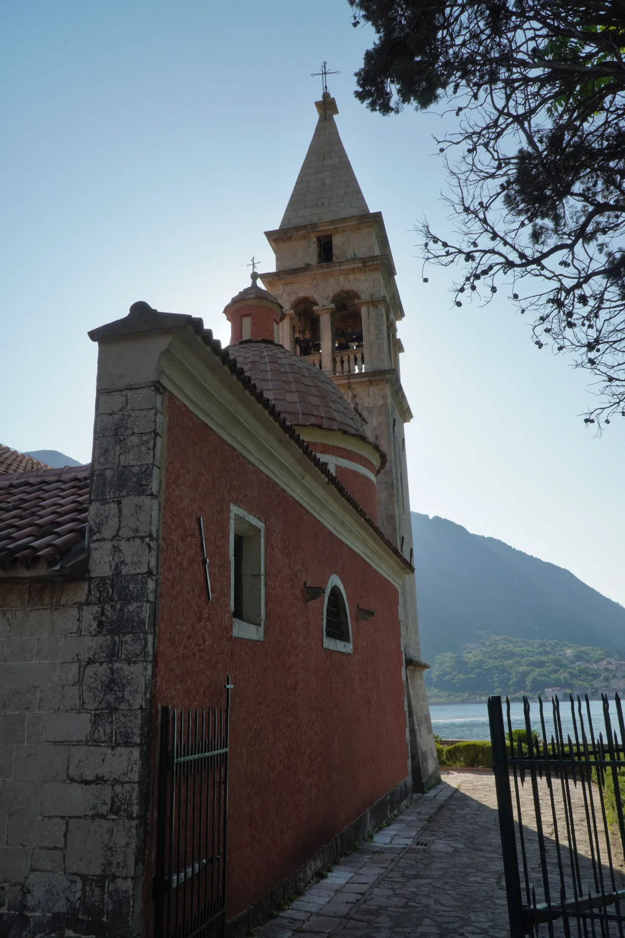 Close-up of a red church building with a tiled dome and a tall stone bell tower reaching towards the sky, overlooking the Bay of Kotor with mountains in the distance.