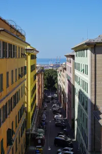 View looking down a narrow street in Genoa, Italy, flanked by tall, colorful buildings. Cars are parked along the street, and in the distance, the port with cranes and the sea are visible under a clear blue sky.