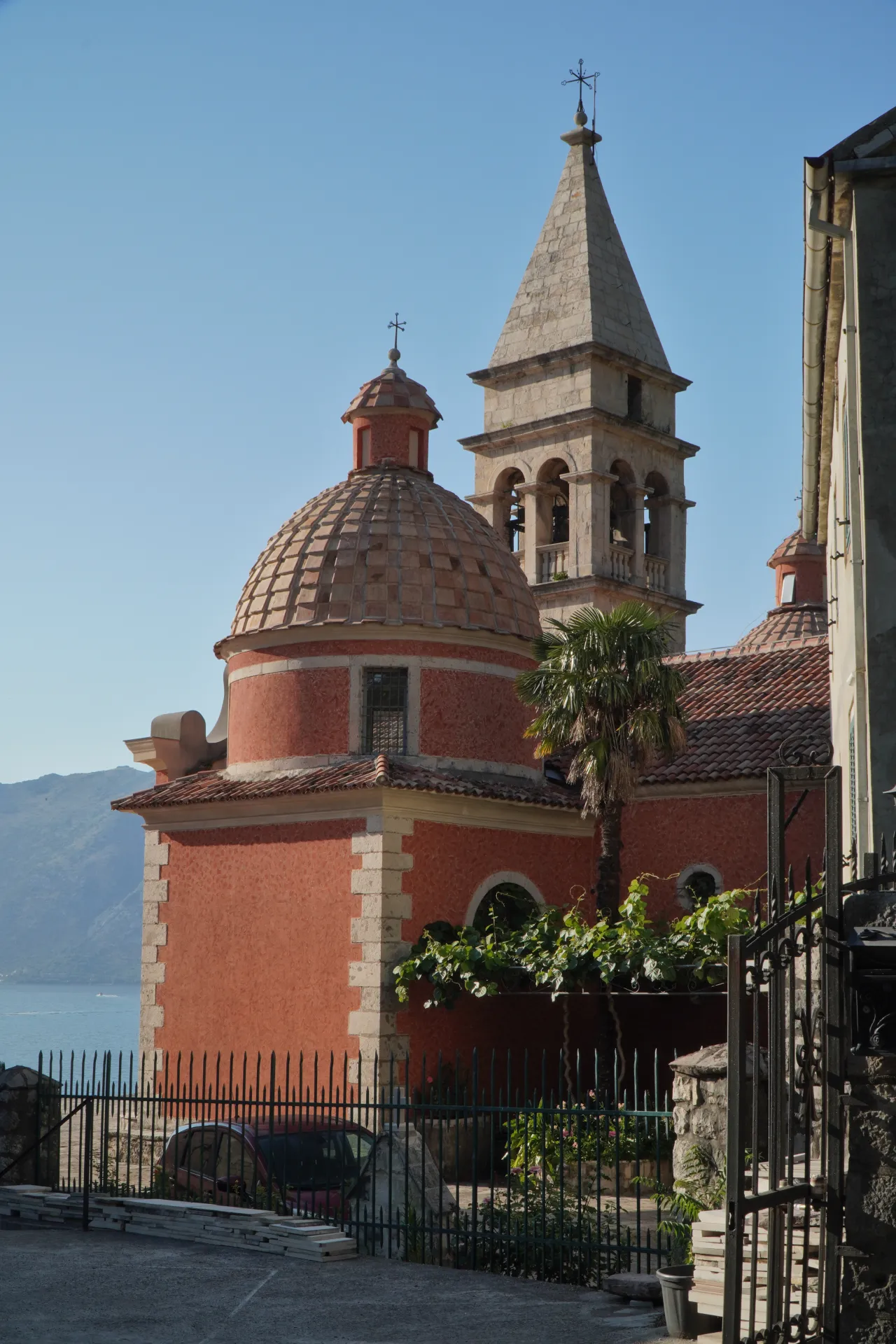 A striking red church with a tiled dome and a stone bell tower stands proudly under a clear blue sky in Kotor, Montenegro, with the calm blue waters of the bay and rolling hills visible in the background.
