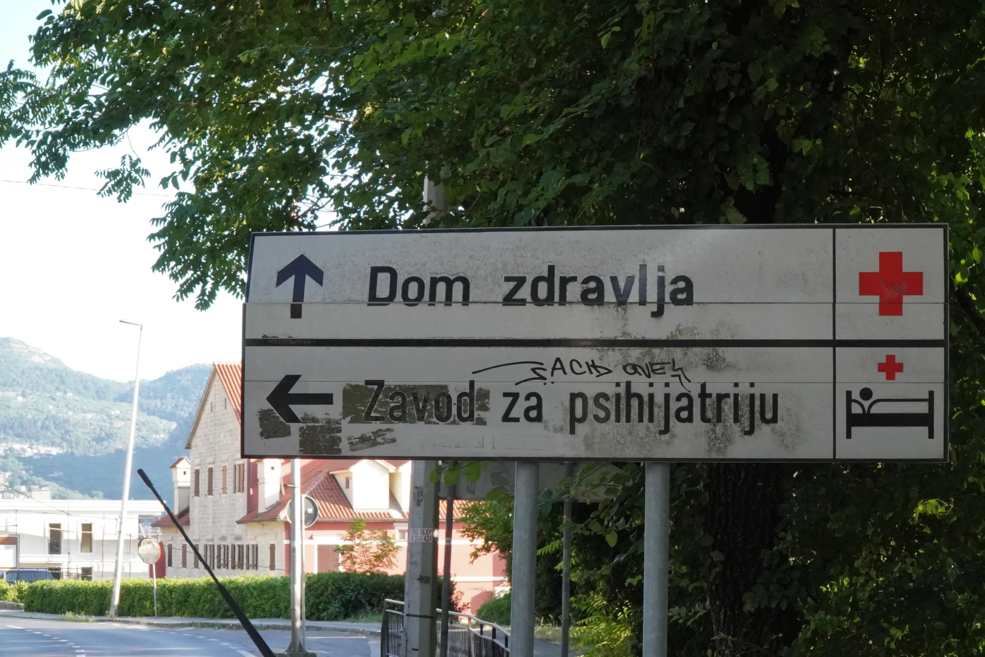 A street sign in Kotor, Montenegro, with two panels. The top panel shows an upward arrow and the text 'Dom zdravlja' (Health Center) with a red cross symbol. The bottom panel shows a leftward arrow and the text 'Zavod za psihijatriju' (Institute of Psychiatry) with a red cross and a bed symbol. Trees and buildings are visible in the background.