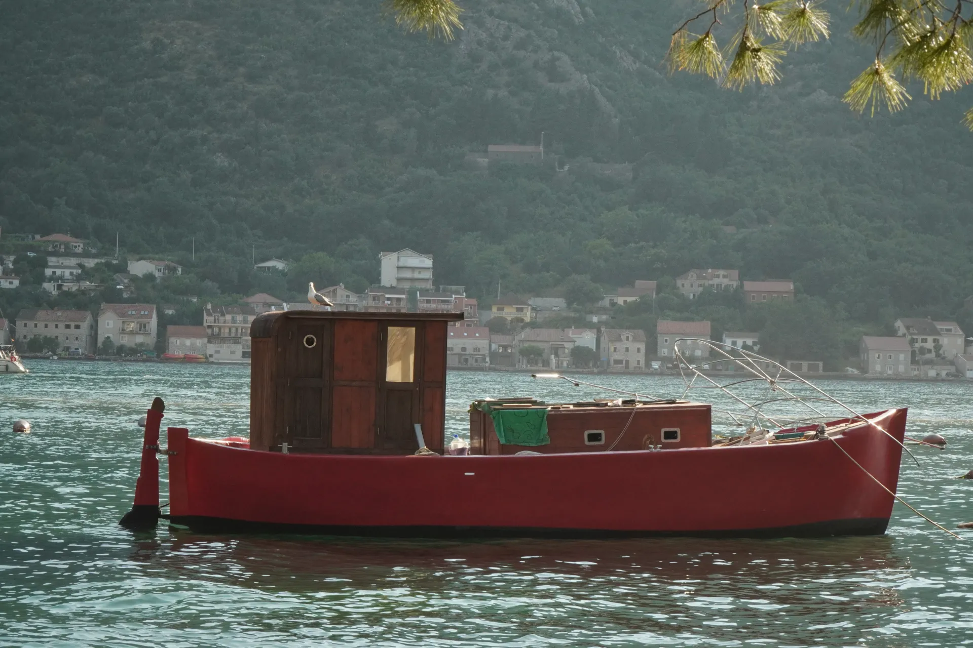 A vibrant red fishing boat with a wooden cabin sits peacefully on the shimmering water of Kotor Bay, Montenegro. In the background, houses are nestled on a verdant, tree-covered mountainside.