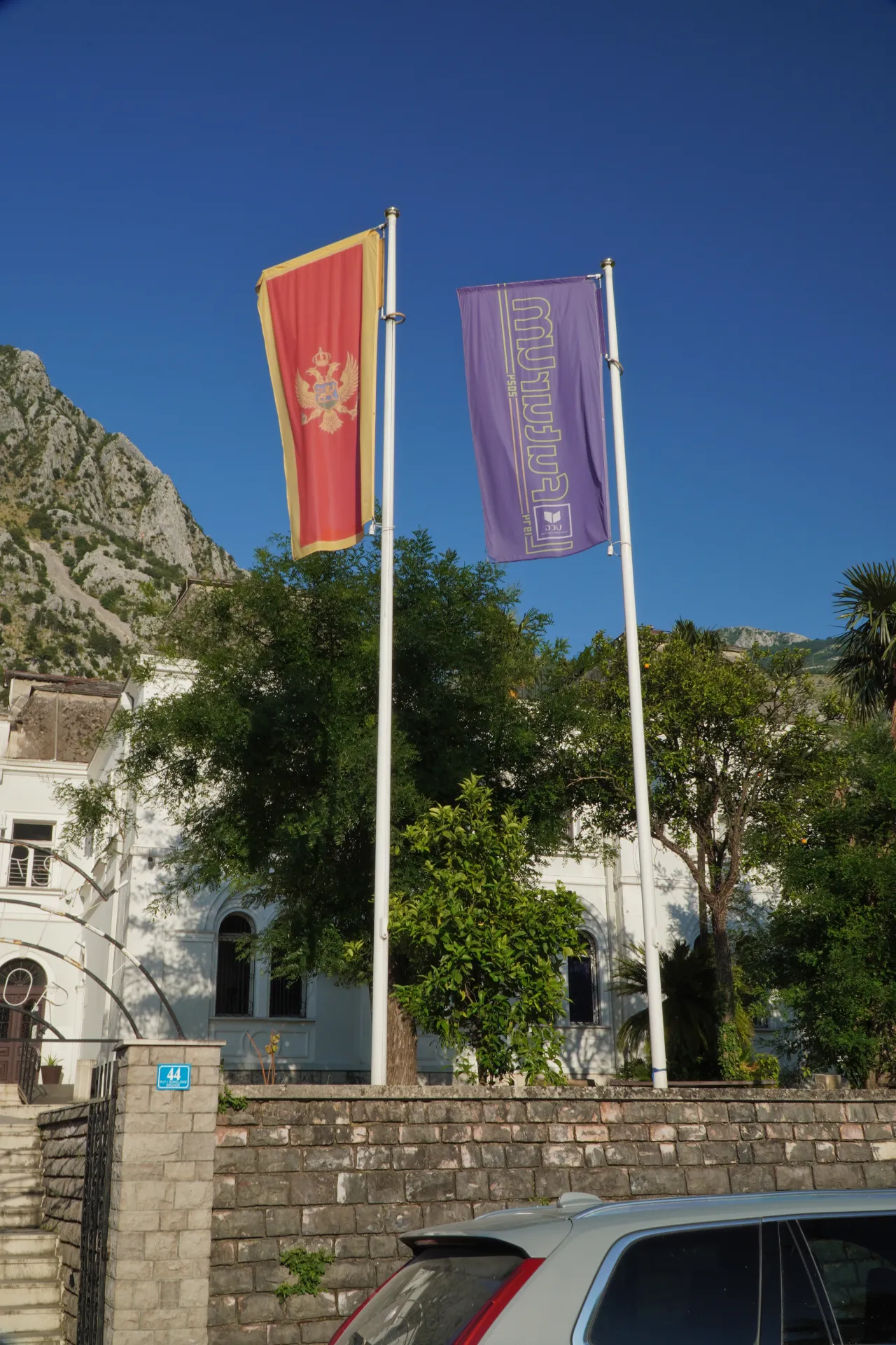 Crnogorska zastava i zastava Pomorskog muzeja Kotor vijore se ispred bele zgrade sa drvecem i planinom u pozadini.