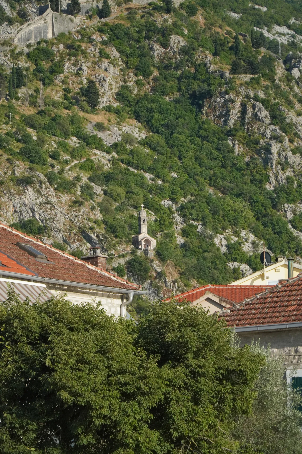 Mala kamena crkva sa zvonikom stoji na zelenoj, stenovitoj planini iznad krovova Kotora u Crnoj Gori prekrivenih crvenim crepom. Drevni bedemi tvrđave vidljivi su više uz padinu.