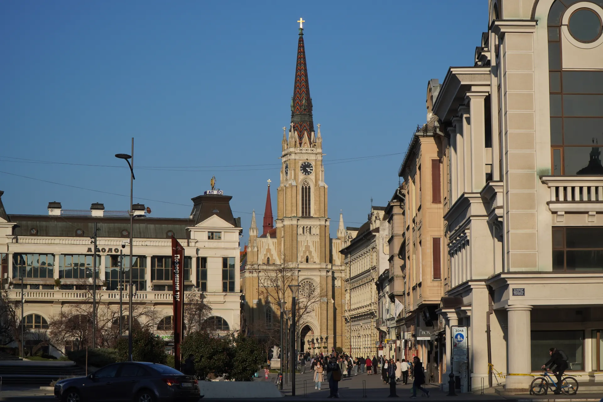The impressive Cathedral of the Name of Mary in Novi Sad, Serbia, with its tall spire, stands against a clear blue sky, surrounded by other historic buildings and a busy street.