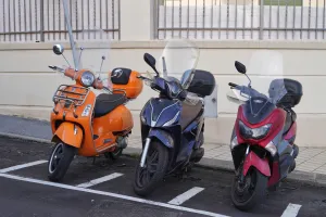 An orange Vespa scooter, a dark blue scooter, and a red scooter are parked in a row on a street with white parking lines.