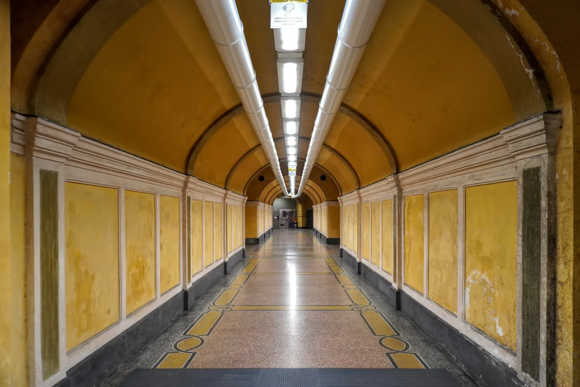 A long, symmetrical tunnel with yellow walls, white trim, a patterned marble floor, and bright ceiling lights.