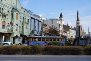 A street in Novi Sad, Serbia, with a classic blue and wooden tram in the foreground, surrounded by a mix of historical and modern buildings under a clear blue sky. The Serbian flag flies from a balcony.