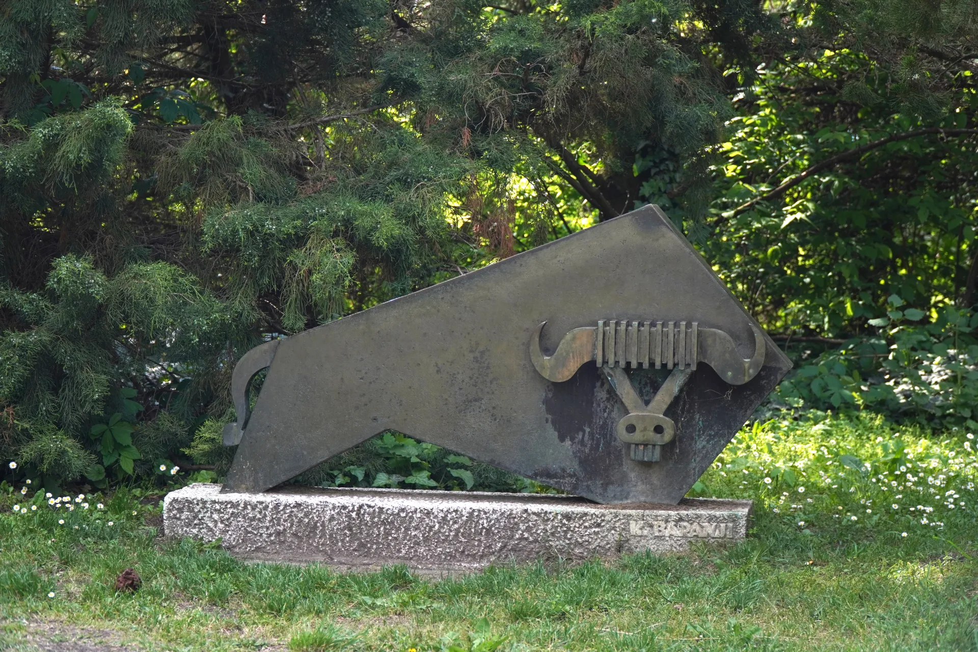 An abstract metal sculpture of a bull's head with prominent horns and a stylized face rests on a stone base in a park, with trees and green foliage in the background.