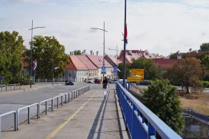 A street in Novi Sad, Serbia, with buildings featuring red tiled roofs, Serbian flags flying from lampposts, and a person walking on a pedestrian bridge.