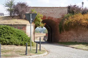 An old, weathered brick archway leading into Petrovaradin Fortress, Novi Sad, Serbia, with green bushes and trees on either side and a view of the city in the distance.