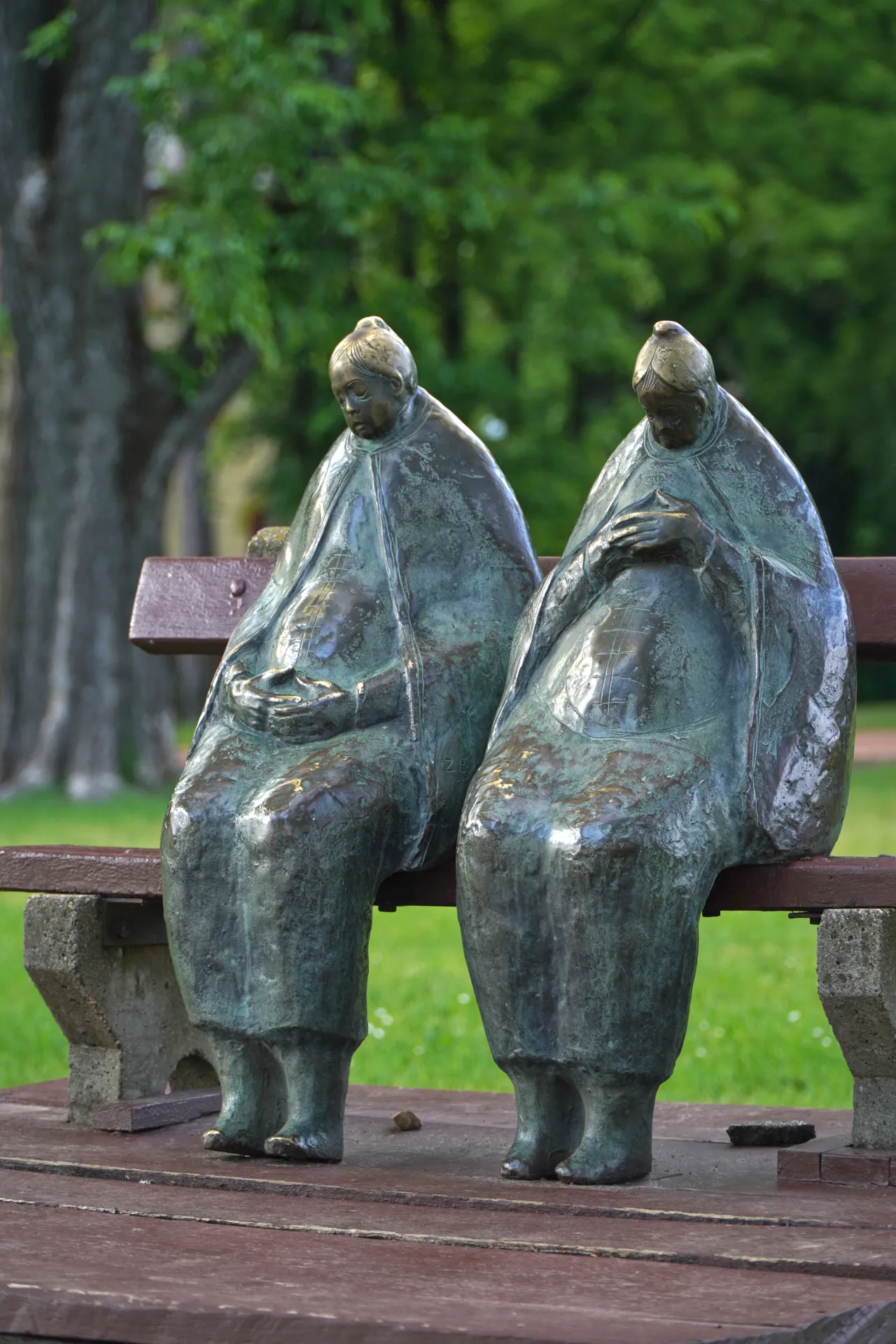 A bronze sculpture of two elderly women in long robes sitting on a wooden park bench in Palic, Serbia, with a green, tree-filled background.