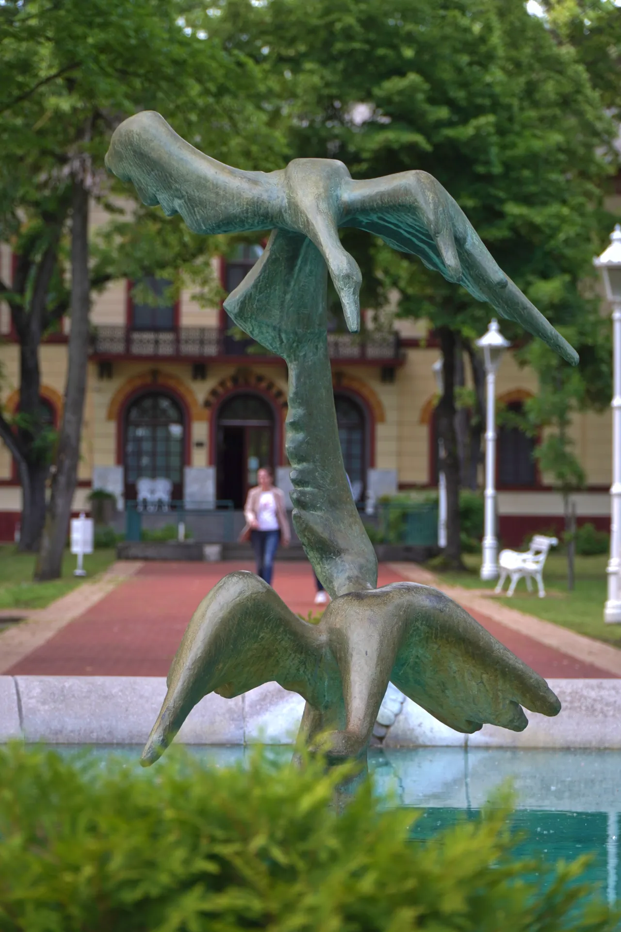 A weathered bronze sculpture of two swans in mid-flight, with a building and trees in the background in Palic, Serbia.