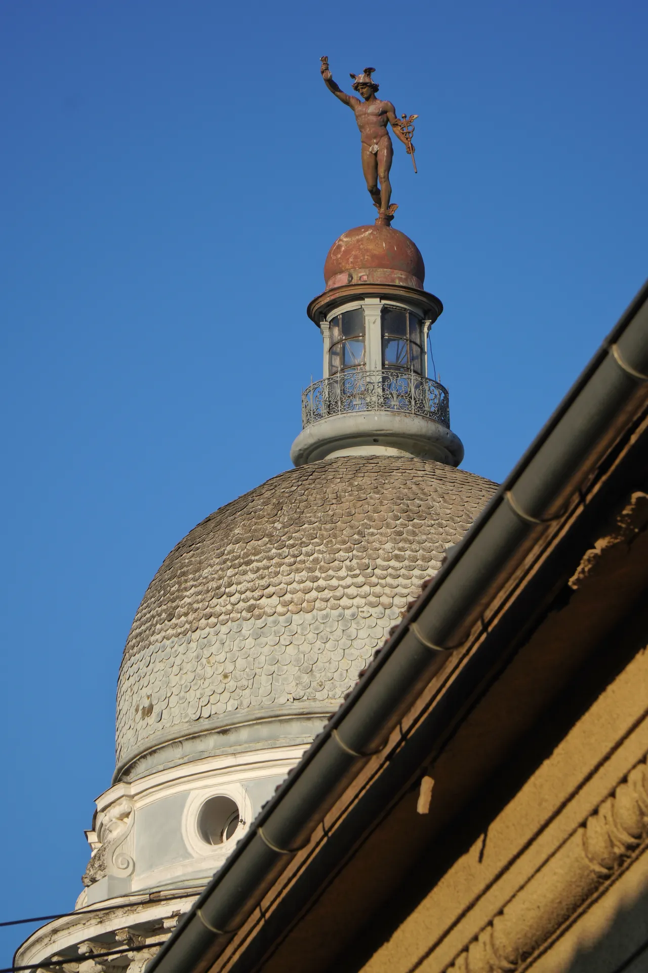A statue of the god Mercury with wings on its helmet and holding a caduceus, stands on top of a copper-domed building with a clear blue sky background in Novi Sad, Serbia.
