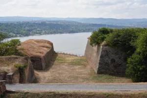 View of the Danube River and the city of Novi Sad from the ramparts of Petrovaradin Fortress, with stone walls and grassy slopes in the foreground and hills in the background.