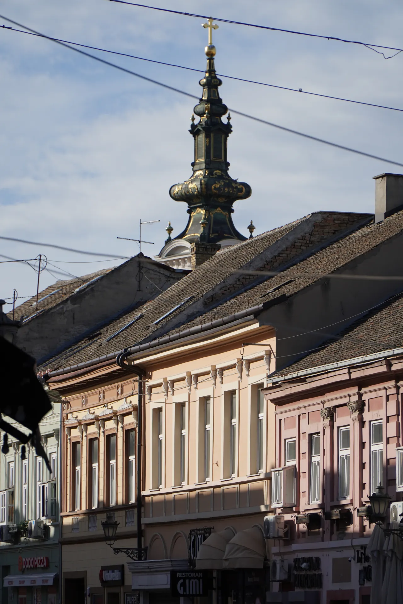 The ornate green and gold spire of the Novi Sad Cathedral is visible above the pitched tile roofs of old buildings, under a cloudy blue sky. Wires crisscross the frame, and pastel-colored building fronts line the street below.