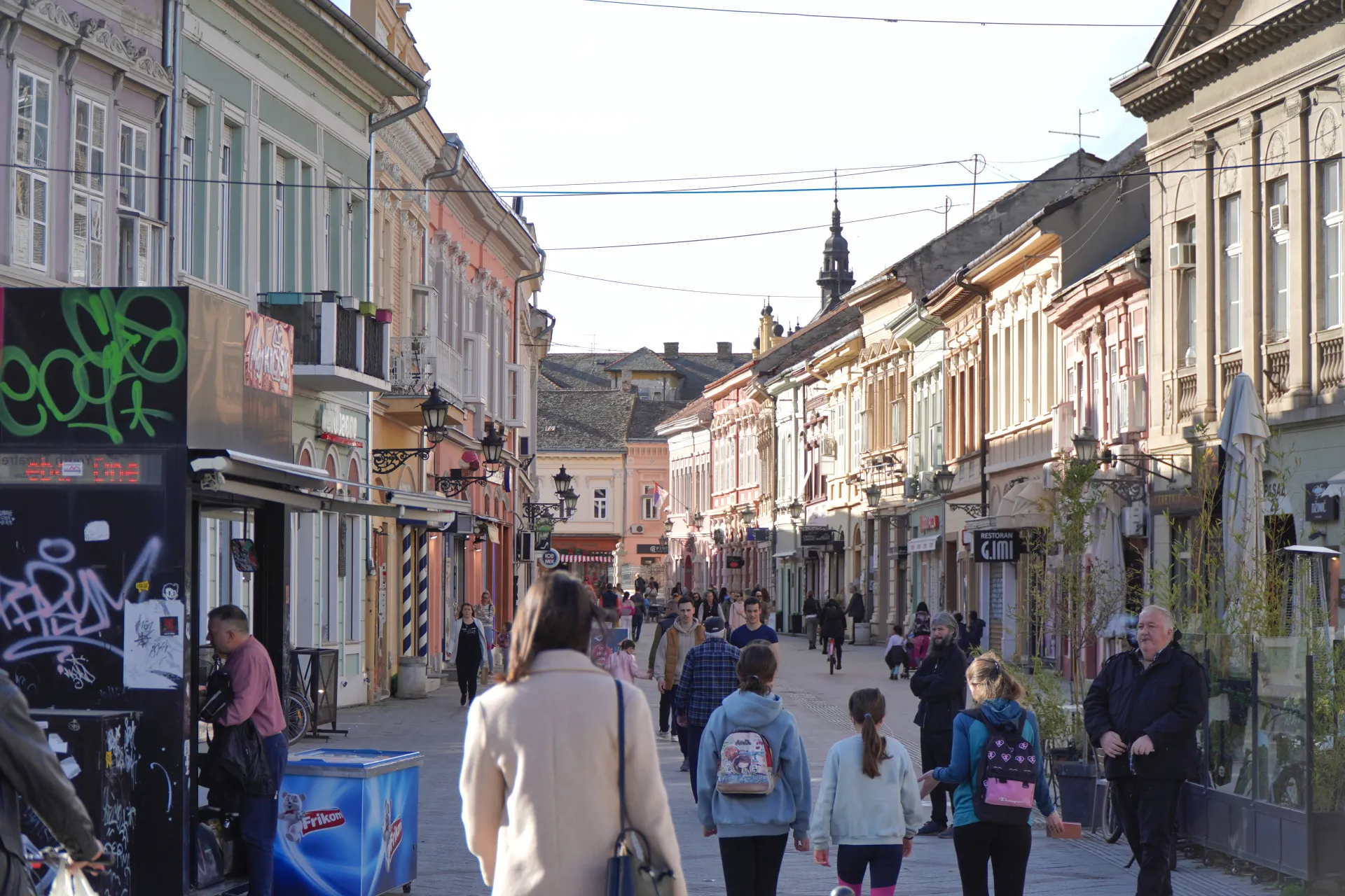 A view down a busy pedestrian street in Novi Sad, Serbia, with colorful buildings, shops, and people walking.