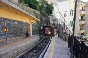 A red funicular train with its headlights on is entering a tunnel on a steep railway track. The view is from the platform of a station which has a yellow roof and is built against a stone wall. To the right, a set of stairs with black railings leads up the side of the incline next to a building with balconies.